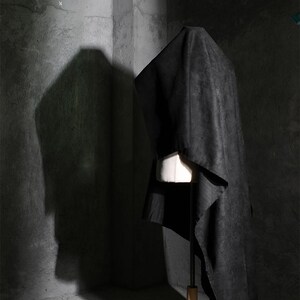 May include: A black garment draped over a white dress form. The fabric has a textured appearance and falls in soft folds. The dress form is on a wooden stand, set against a textured gray wall.