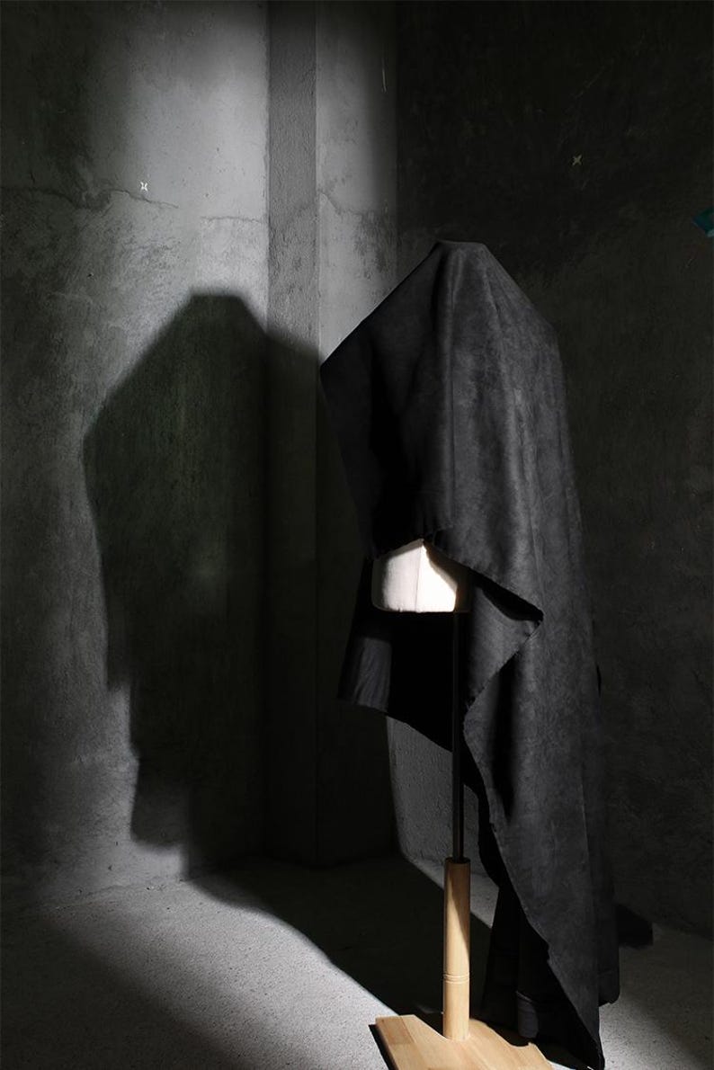 May include: A black garment draped over a white dress form. The fabric has a textured appearance and falls in soft folds. The dress form is on a wooden stand, set against a textured gray wall.