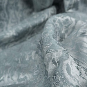 Puede incluir: Primer plano de una tela fluida en tonos azules y blancos. El material tiene un patrón arremolinado y veteado, creando una sensación de movimiento y profundidad. La tela parece suave y lujosa, con sutiles reflejos y sombras.