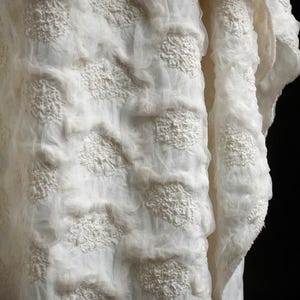 May include: Close-up of a white fabric with a textured, layered design. The fabric features raised, floral-like patterns and a sheer, gauzy texture. The overall effect is soft and delicate, with a slightly frayed edge.