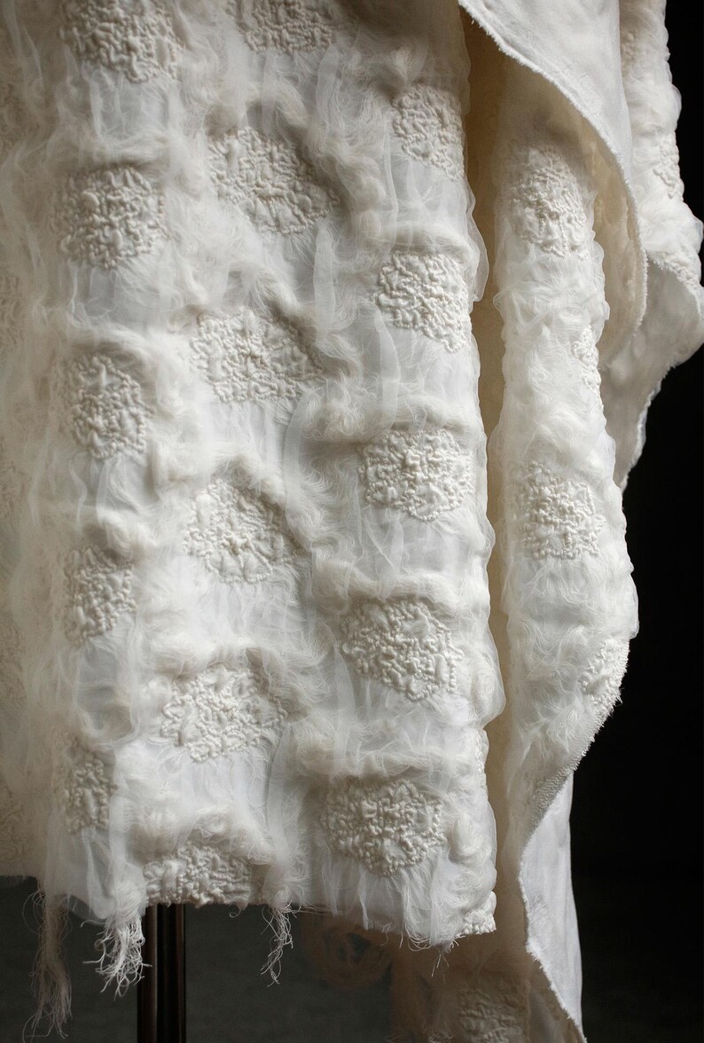 May include: Close-up of a white fabric with a textured, layered design. The fabric features raised, floral-like patterns and a sheer, gauzy texture. The overall effect is soft and delicate, with a slightly frayed edge.
