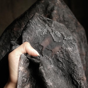 May include: Close-up of a dark, textured fabric with a distressed appearance. The fabric is held by a hand, revealing its intricate surface and subtle color variations. The overall aesthetic suggests a vintage or gothic theme.