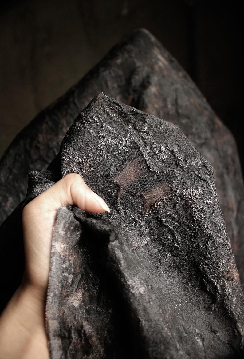 May include: Close-up of a dark, textured fabric with a distressed appearance. The fabric is held by a hand, revealing its intricate surface and subtle color variations. The overall aesthetic suggests a vintage or gothic theme.