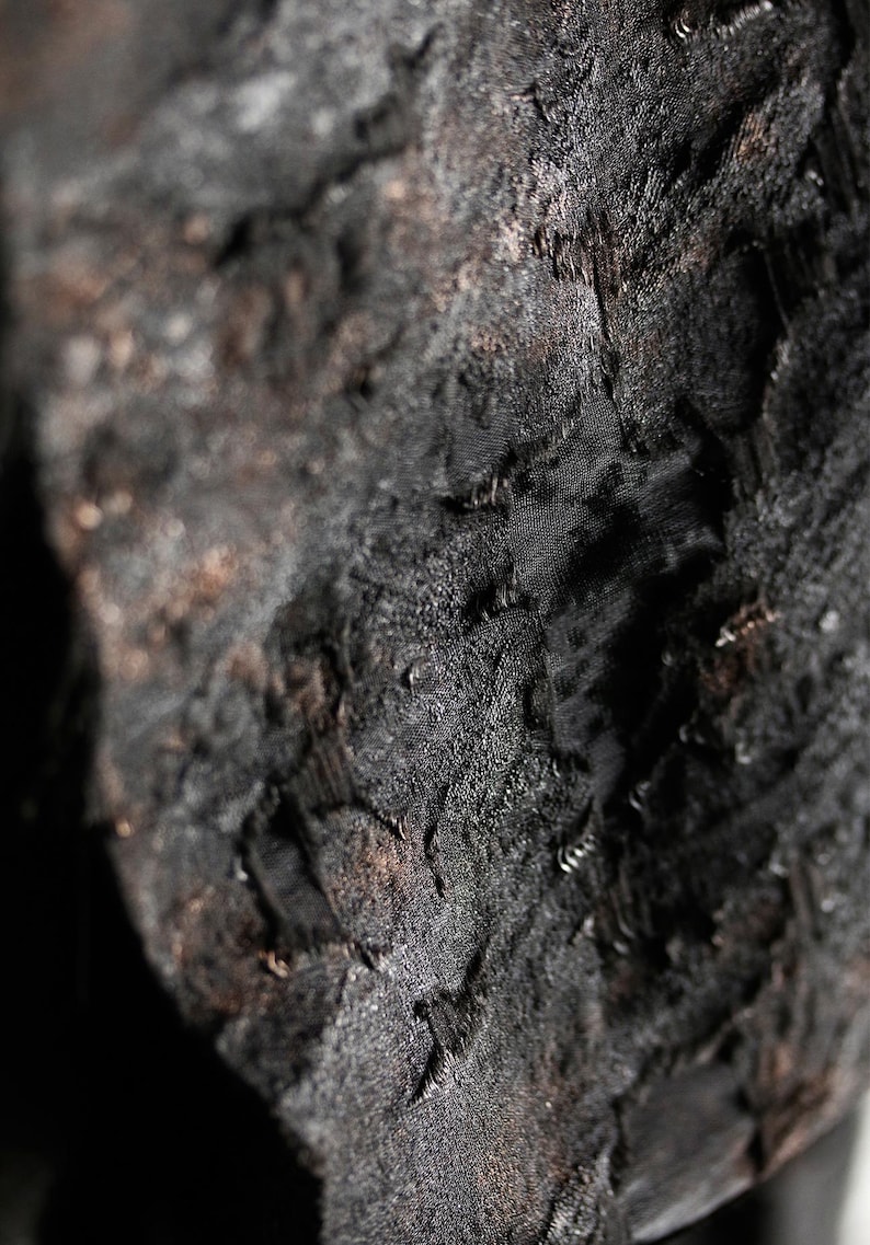 May include: Close-up of a textured, dark charcoal-colored fabric. The surface has a rough, uneven appearance with visible ridges and subtle variations in tone. The fabric appears to be layered, creating depth and shadow.