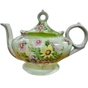 May include: A vintage porcelain teapot with a floral design. The teapot features a light green base and lid, with hand-painted flowers in shades of pink, yellow, and white. The handle and spout are a light gray color.