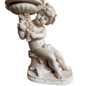 May include: An off-white, cream-colored statue of a cherub sitting on a pedestal base. The cherub holds a shallow, fluted bowl above its head with both hands. The figure features detailed facial expressions, curly hair, and a draped cloth.