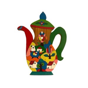 Peut inclure: Une théière en bois colorée avec une poignée verte et un motif peint fantaisiste représentant des maisons, des fleurs et des oiseaux.