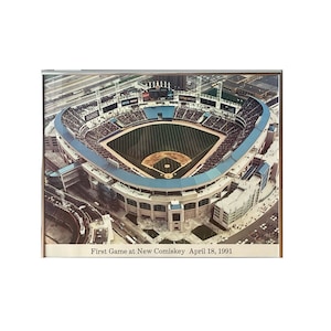 May include: An aerial view of a baseball stadium with a green field and blue seats. The stadium is surrounded by a parking lot and city buildings. The text "First Game at New Comiskey April 18, 1991" is printed below the image.