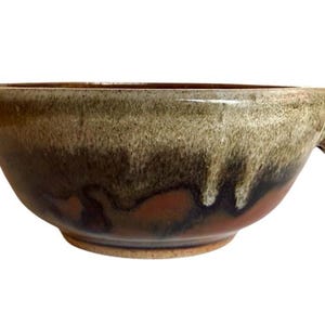 May include: A handmade ceramic bowl with a unique glaze. The bowl features a rounded shape with two small handles and a color gradient of brown, green, and black. The bowl is suitable for serving soups or stews.