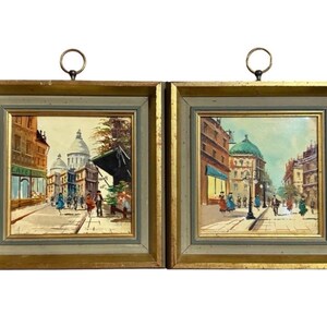 May include: Two framed oil paintings depicting Parisian street scenes. Each painting features a gold frame with a light green inner border and a metal ring for hanging. The artwork showcases buildings, people, and a domed structure.