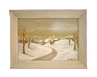 May include: A framed painting of a winter landscape. The artwork depicts a snow-covered scene with a winding road leading to a church. Bare trees and rolling hills are visible under a muted sky. The frame is a light, neutral color.