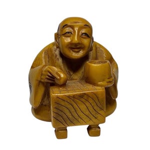 May include: A carved ochre-colored figurine depicting a bald person with a smiling expression, seated behind a rectangular game board with wavy line patterns. The figure holds a small cup in one hand and rests the other hand on the game board surface.