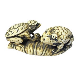 May include: A carved off-white netsuke figurine featuring two textured toads resting on a curved, ribbed leaf. The toad on the left wears a small flower-shaped hat, while the toad on the right has dark, detailed markings around its eyes and back.