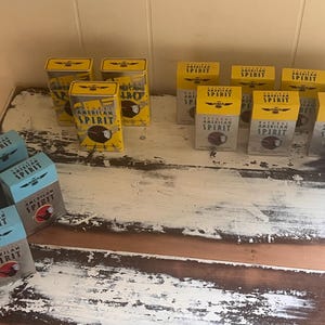 May include: Assortment of Natural American Spirit cigarette boxes and tins. The boxes are primarily yellow and gray, with the brand name and logo prominently displayed. The tins are yellow with a clear plastic window. The boxes are blue and gray.