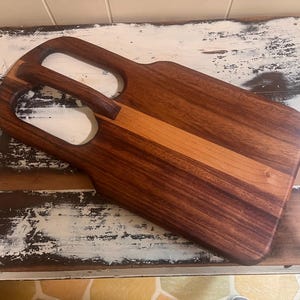 Peut inclure: Une planche à découper en bois avec une poignée. La planche est rectangulaire avec des coins arrondis et une bande marron clair au centre. La poignée comporte deux ouvertures pour faciliter le transport. Le grain du bois est visible.