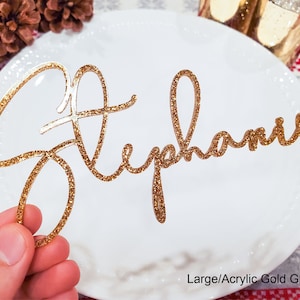 May include: Gold glitter acrylic name card with the name "Stephanie" written in cursive script. The card is on a white plate with a red and white checkered tablecloth in the background.