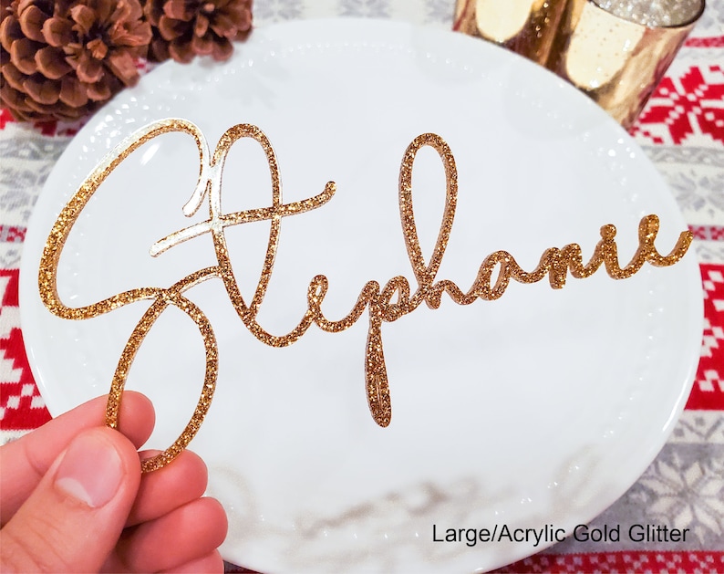 May include: Gold glitter acrylic name card with the name "Stephanie" written in cursive script. The card is on a white plate with a red and white checkered tablecloth in the background.