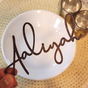 May include: A wooden name cutout spelling "Aaliyah" in a cursive font. The dark brown cutout is held in front of a white bowl and two glasses. The background has a gold, textured surface.