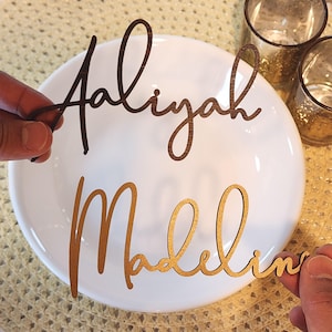 May include: Two wooden name cutouts, "Aaliyah" in dark brown and "Madeline" in gold, are held above a white plate. The names are in a cursive font. The background includes two small glass candle holders and a textured surface.