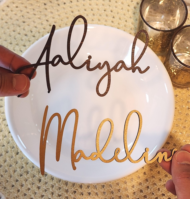 May include: Two wooden name cutouts, "Aaliyah" in dark brown and "Madeline" in gold, are held above a white plate. The names are in a cursive font. The background includes two small glass candle holders and a textured surface.