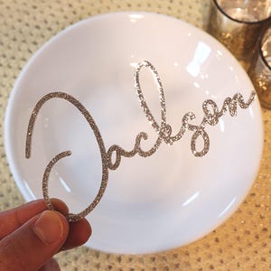 May include: A silver glitter "Jackson" name cutout in cursive script is held above a white bowl. The name is made of sparkling material. The bowl is white and glossy. The background is a textured, golden surface.