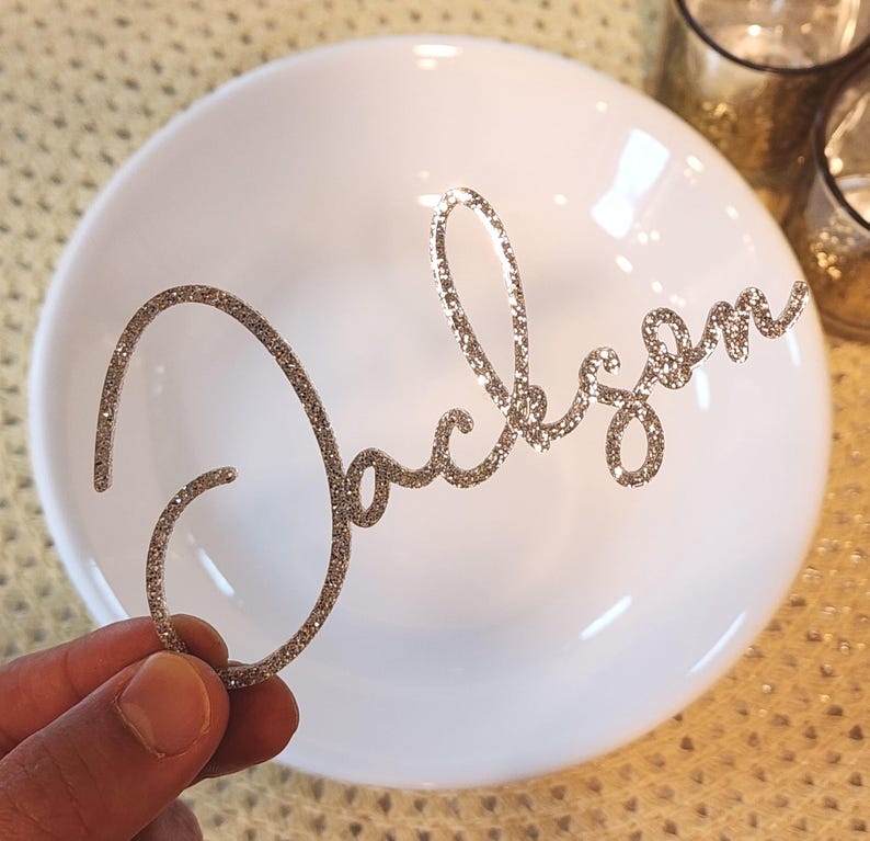 May include: A silver glitter "Jackson" name cutout in cursive script is held above a white bowl. The name is made of sparkling material. The bowl is white and glossy. The background is a textured, golden surface.