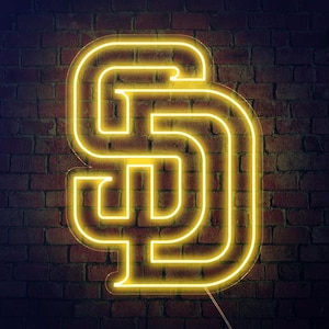 Pode incluir: Uma placa de néon amarela brilhante com as letras "SD" numa parede de tijolos escuros. O design luminoso e estilizado cria um contraste marcante.