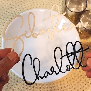 May include: Two laser-cut name place cards. One is a light beige colour and reads "Sebastian", the other is black and reads "Charlott". Both names are in a cursive font. The place cards are held in front of a white plate.