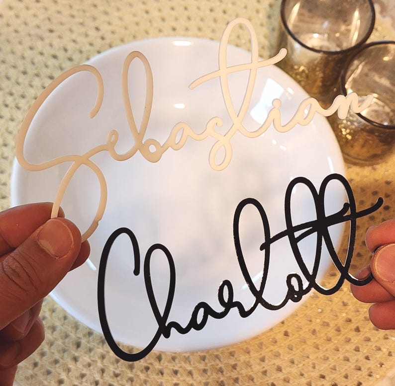 May include: Two laser-cut name place cards. One is a light beige colour and reads "Sebastian", the other is black and reads "Charlott". Both names are in a cursive font. The place cards are held in front of a white plate.