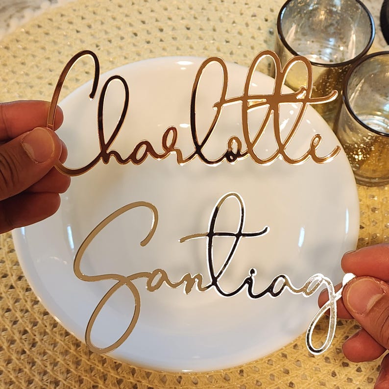 May include: Gold and silver laser-cut name place cards. The names "Charlotte" and "Santiago" are displayed in elegant cursive script. The place cards are held above a white plate, suggesting a table setting for a special event.