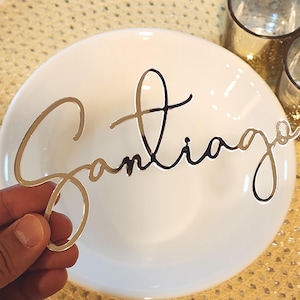 May include: A gold-coloured metallic name cutout spelling "Santiago" in a cursive font. The cutout is held in front of a white plate. Two small glass candle holders are in the background.