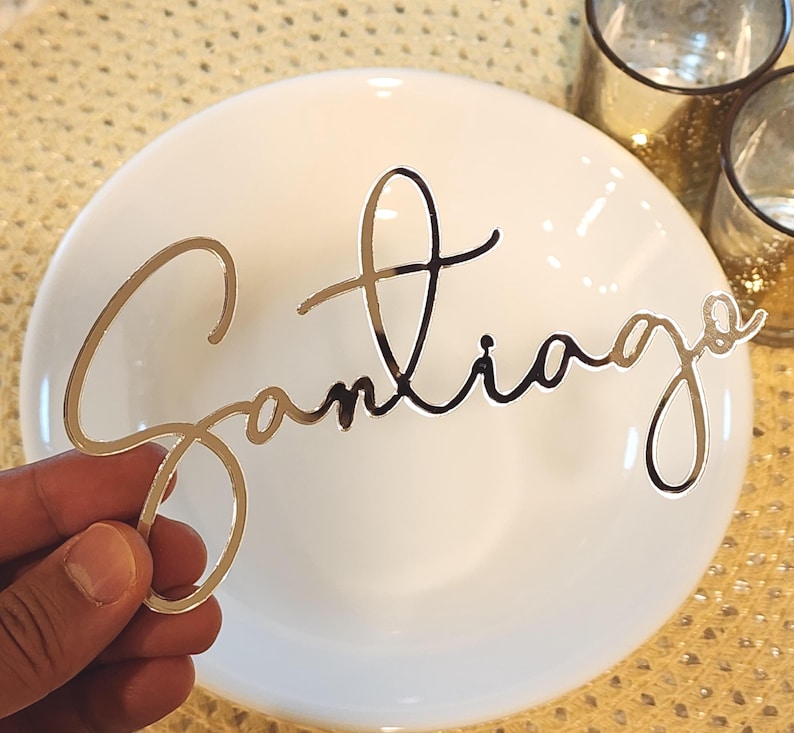 May include: A gold-coloured metallic name cutout spelling "Santiago" in a cursive font. The cutout is held in front of a white plate. Two small glass candle holders are in the background.