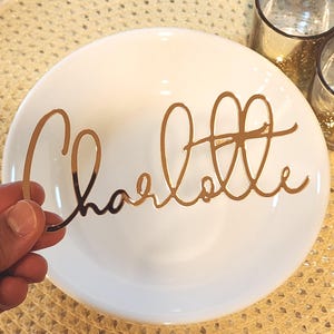 May include: A gold-coloured, cursive-style nameplate reading "Charlotte" is held in front of a white plate. The nameplate is made of a reflective material. The plate is on a textured, gold-coloured surface.