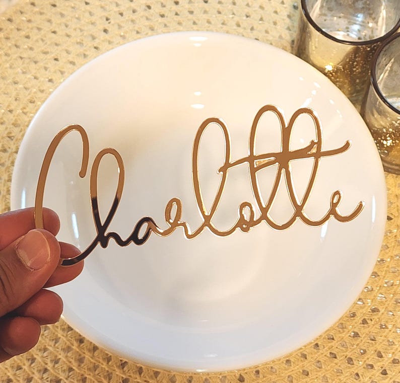May include: A gold-coloured, cursive-style nameplate reading "Charlotte" is held in front of a white plate. The nameplate is made of a reflective material. The plate is on a textured, gold-coloured surface.