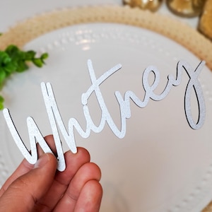 May include: A silver-colored wooden name cutout that spells "Whitney" in a cursive font. The cutout is held in front of a white plate and a beige placemat. Decorative elements are in the background.