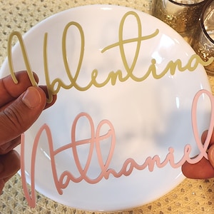 May include: Two cursive name cutouts, "Valentina" in gold and "Nathaniel" in pink, are held over a white plate. The names are in a delicate, flowing script, ideal for personalised event decor or gifts.