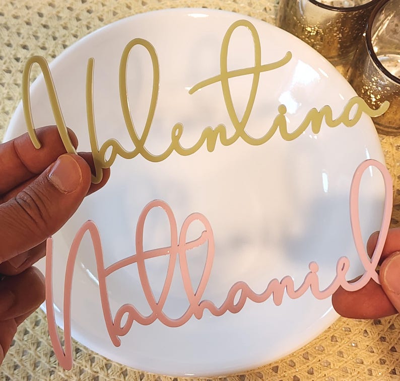 May include: Two cursive name cutouts, "Valentina" in gold and "Nathaniel" in pink, are held over a white plate. The names are in a delicate, flowing script, ideal for personalised event decor or gifts.