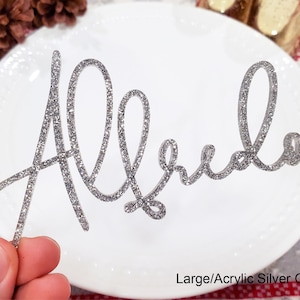 May include: Silver glitter acrylic name card with the name "Alfredo" written in cursive script. The card is on a white plate with a red and white patterned surface in the background.