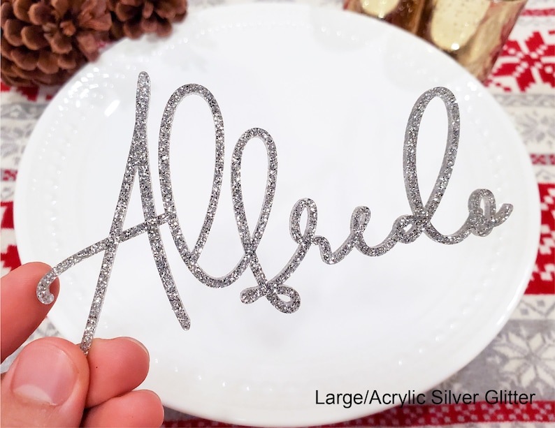 May include: Silver glitter acrylic name card with the name "Alfredo" written in cursive script. The card is on a white plate with a red and white patterned surface in the background.