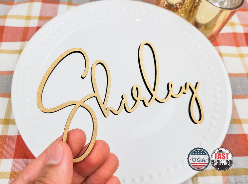 May include: A gold wooden name place card with the name "Shirley" written in a cursive script.