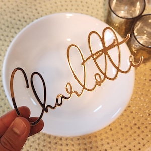 May include: A gold-coloured, cursive nameplate reading "Charlotte" is held above a white plate. The nameplate is made of a shiny, reflective material. Two small, clear glasses are in the background.