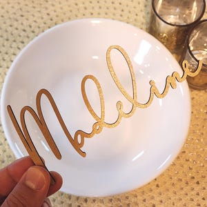 May include: A gold-coloured wooden name cutout spelling "Madeline" in cursive. The cutout is held in front of a white plate, possibly for a place setting. The background features a textured surface and glassware, suggesting a dining setting.