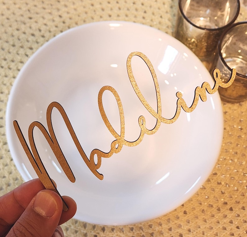 May include: A gold-coloured wooden name cutout spelling "Madeline" in cursive. The cutout is held in front of a white plate, possibly for a place setting. The background features a textured surface and glassware, suggesting a dining setting.