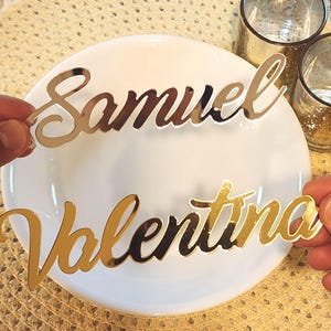 May include: Two metallic name place cards, one reading "Samuel" in a silver finish and the other "Valentina" in gold. The cards are held above a white plate, suggesting a table setting for an event.
