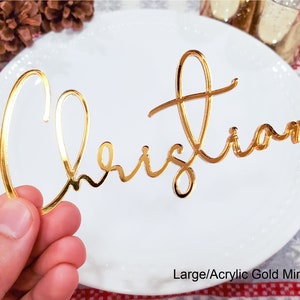May include: Gold mirror acrylic place card with the name "Christian" written in a cursive script.