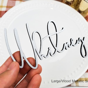 May include: A silver metallic wood place card with the name "Whitney" written in a cursive font.