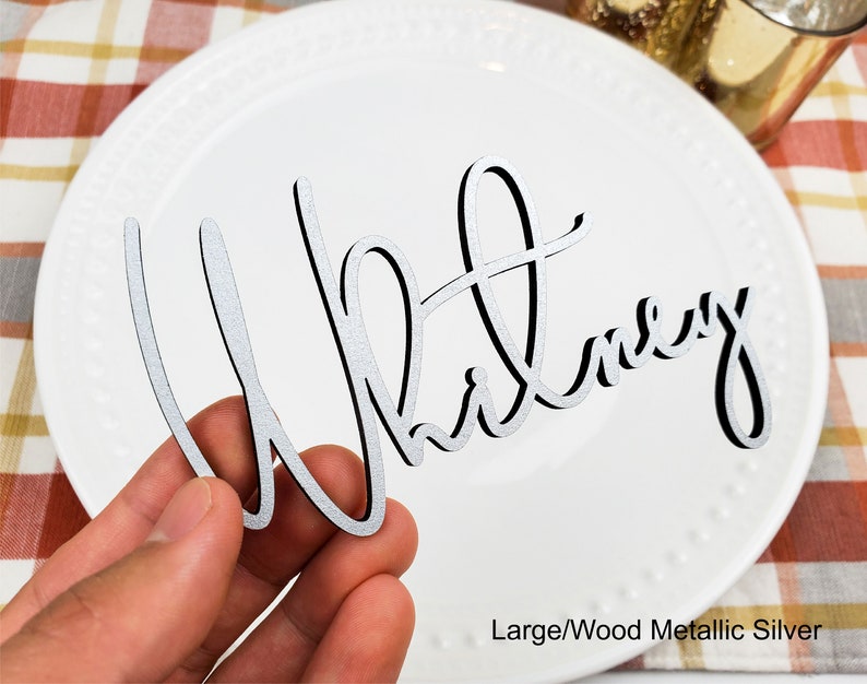May include: A silver metallic wood place card with the name "Whitney" written in a cursive font.