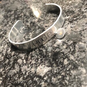 Puede incluir: Una pulsera de puño plateada con la palabra "Pride" grabada. Un pequeño dije redondo con un corazón dorado cuelga de la pulsera. La pulsera se muestra sobre una superficie gris moteada.
