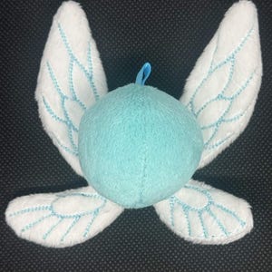 May include: A plush fairy doll with a light blue body and white wings. The wings are detailed with light blue stitching, and the doll has a small blue loop at the top. The fairy is designed to look like a cartoon character.