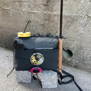 May include: A black fly fishing backpack with a yellow fly reel and a brown fly rod. The backpack has a mesh pocket and a zipped pocket. The fly rod is leaning against the backpack.
